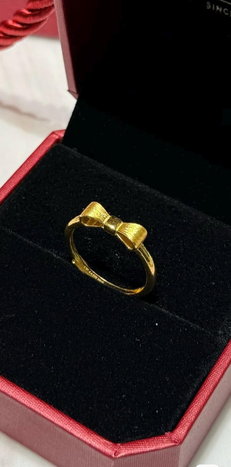 A gold ring with a bow design is displayed in a red jewelry box.