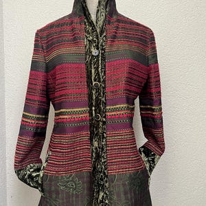 Può includere: Una giacca a maniche lunghe con un motivo a righe orizzontali multicolori nei toni del rosso, viola e verde. Il capo presenta un colletto alto, una chiusura frontale con bottoni e un bordo in velluto verde scuro lungo l'abbottonatura.