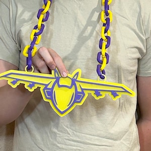 May include: A yellow and purple airplane-shaped pendant necklace. The pendant has a yellow center with purple outlines and wings. The chain is made of alternating yellow and purple links. The necklace is being held up.