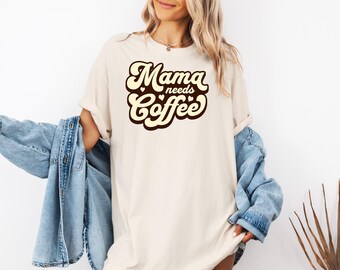 May include: A white t-shirt with the text "Mama needs coffee" in brown and cream retro lettering.