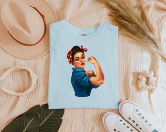 May include: Light blue t-shirt with a graphic of a woman with red and white polka dot headband flexing her bicep. The woman is wearing a blue shirt and has brown hair.