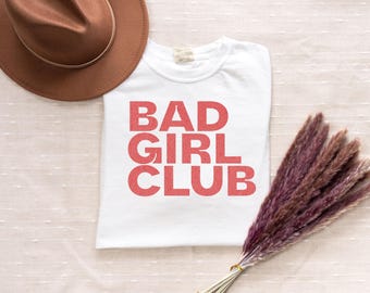 May include: White t-shirt with the text "BAD GIRL CLUB" printed in pink.
