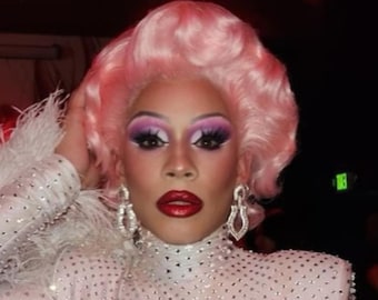 May include: A person wearing a white sequined top with a pink wig and large diamond earrings. The person has on bold makeup with red lipstick.