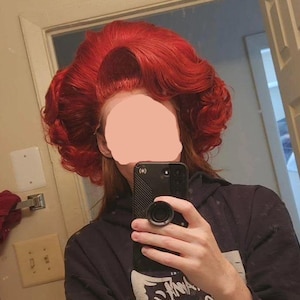 May include: A person with vibrant red hair styled in a voluminous, vintage-inspired updo. The hair is a bold, saturated color, with curls and waves creating a dramatic silhouette. The person is wearing a black hoodie and holding a black smartphone.
