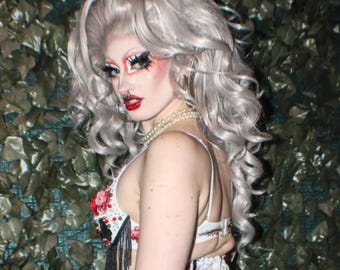May include: A person with silver-gray hair and dramatic makeup, including bold eye shadow and red lipstick. They wear a white corset-style top with floral and fringe details, a pearl necklace, and a nose ring. The background features green foliage.