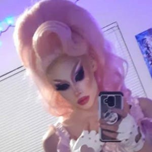 May include: A person with dramatic makeup and a pink wig is holding a phone, taking a selfie. They are wearing a light pink outfit with white gloves that have heart-shaped cutouts. The background is a purple hue.