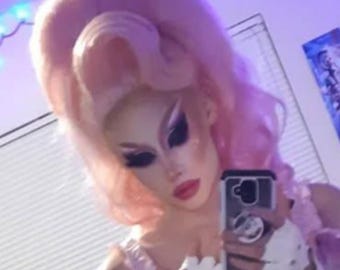 May include: A person with dramatic makeup and a pink wig is holding a phone, taking a selfie. They are wearing a light pink outfit with white gloves that have heart-shaped cutouts. The background is a purple hue.