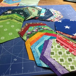 May include: A selection of patterned fabric pieces in shades of green, blue, red, and purple, arranged on a blue cutting mat. These fabric pieces are ideal for quilting or other craft projects.