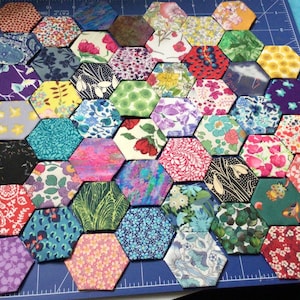 May include: A collection of colourful fabric hexagons arranged in a honeycomb pattern on a blue cutting mat. The hexagons are made of various floral prints and patterns.