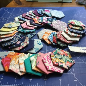 May include: A collection of colourful fabric hexagons, some with floral patterns, arranged in a circular pattern on a blue cutting mat. The label "LIBERTY FABRICS" is visible in the bottom right corner.