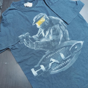 May include: Dark blue t-shirt featuring a detailed graphic of a futuristic soldier holding a weapon. The design includes the text "HALO ANNIVERSARY" and "COMBAT EVOLVED" in white. The shirt is made of a soft, casual fabric.
