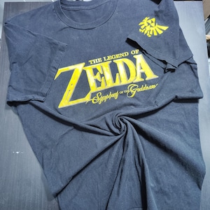May include: Dark gray t-shirt featuring the text "THE LEGEND OF ZELDA Symphony of the Goddesses" in yellow. A yellow Triforce logo is on the right sleeve. The shirt is made of a soft, comfortable material.