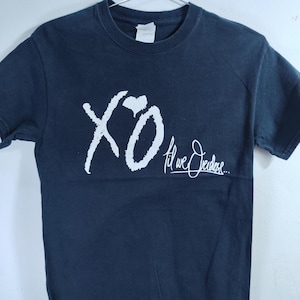 May include: Navy blue t-shirt with a white "XO" graphic and cursive text. The short-sleeved shirt has a crew neck and is made of a soft material. The graphic design is located on the front of the shirt.