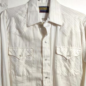 Może przedstawiać: Kremowa koszula z długim rękawem i pionowymi prążkami. Posiada dwie przednie kieszenie z zapięciem na guziki i spiczasty kołnierz. Koszula ma karczek w stylu western i etykietę z napisem "Wrangler Slim".