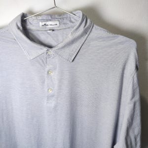 以下が含まれることがあります： 襟と3つのボタンが付いた、水色と白のストライプのポロシャツ。シャツは金属製のハンガーに吊るされています。「PETER MILLAR」のブランド名が襟に表示されています。