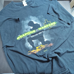 May include: Dark gray t-shirt featuring a kneeling soldier silhouette with the text "THE WEAPONS OF OUR WARFARE ARE NOT CARNAL... CALLED TO DUTY". The design includes a cross and a green and yellow color scheme.