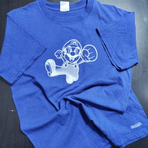 May include: A blue t-shirt featuring a white graphic of the video game character Mario in a dynamic pose. The shirt has short sleeves and a crew neck. The Nintendo logo is visible on the sleeve.