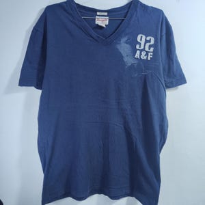 May include: A navy blue V-neck t-shirt with short sleeves. The shirt has the number "92" and the letters "A&F" printed on the front. A small tag is visible near the neckline.