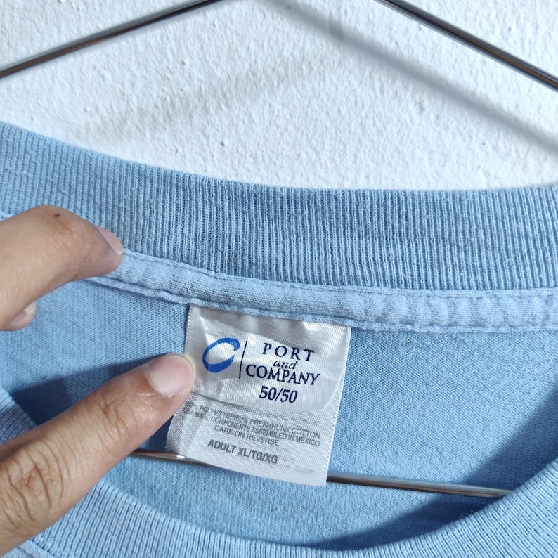 Może przedstawiać: Jasnoniebieska koszulka z metką Port and Company. Na metce widnieje napis "50/50" i "ADULT XL/TG/XG". Koszulka wykonana jest z mieszanki bawełny i poliestru.