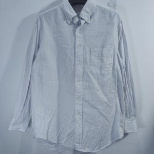 May include: A long-sleeve button-down shirt in a solid white color. The shirt features a button-down collar, a single chest pocket on the left side, and a full-length front button placket with matching white buttons on the cuffs and front.