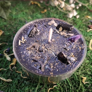 Könnte beinhalten: Eine schwarze Teelichtkerze mit goldenem Glitzer, schwarzen und violetten Kristallen und getrockneten Kräutern.