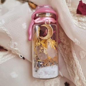 Könnte beinhalten: Kleine Glasflasche gefüllt mit Schichten aus Zutaten, darunter Kristalle, getrocknete Blumen und Sand. Die Flasche ist mit rosa Wachs und einem goldenen Halbmond-Anhänger versiegelt. Der Hintergrund ist ein weicher, cremefarbener Stoff mit Spitzendetails.
