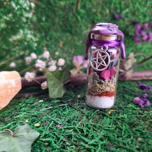 Könnte beinhalten: Eine kleine Glasflasche gefüllt mit Schichten farbenfroher Zutaten, darunter weiße, rote und grüne Elemente. Die Flasche ist mit violettem Wachs und einem silbernen Pentagramm-Anhänger versiegelt. Der Hintergrund zeigt grünes Moos und Blätter.