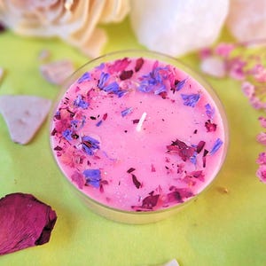 Könnte beinhalten: Eine rosa Kerze in einem transparenten Glasbehälter, verziert mit getrockneten Blütenblättern in Lila, Rosa und Rot. Die Kerze steht auf einer hellgrünen Oberfläche, umgeben von dekorativen Elementen wie Steinen und Trockenblumen. Ein einzelner weißer Docht ist sichtbar.