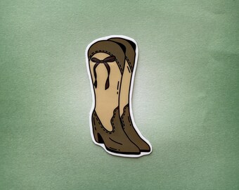 Op de afbeelding: Een sticker met een paar bruine en beige cowboylaarzen. De laarzen hebben een decoratieve strik aan de bovenkant en een geschulpte rand. De achtergrond is effen groen.