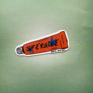 May include: Orange sticker in the shape of a tube of Super Glue. The word "SUPERGLUE" is written in black, with two blue star accents. The words "ROLE MODEL" are written in black at the bottom.