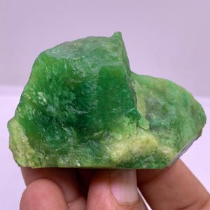 Pode incluir: Um pedaço bruto e não cortado de jade verde. O jade tem uma cor verde vibrante e rica com veios verdes e brancos mais claros. A pedra é segurada em uma mão, mostrando sua forma e textura natural e irregular. Esta pedra preciosa é ideal para fazer joias ou colecionar.