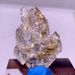 Puede incluir: Un grupo de cristales de cuarzo transparente con múltiples puntas e inclusiones naturales. El cristal está montado sobre una pequeña base azul. El cuarzo tiene un aspecto translúcido con algunas zonas de coloración marrón dorada.