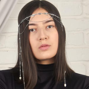 May include: A silver-toned headpiece with delicate chains and small pearl accents. The headpiece is worn on the head and drapes down the sides. The model is wearing a black turtleneck top.