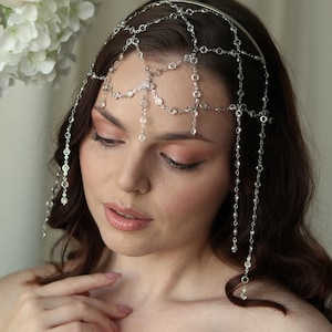 May include: A silver headpiece with dangling crystal details. The headpiece has a delicate chain design with multiple strands of clear, sparkling crystals. The headpiece is worn on the head and drapes down the forehead and sides.
