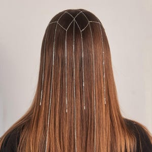 以下が含まれることがあります： 幾何学模様のシルバーヘッドチェーンと垂れ下がるチェーン。長い茶色の髪に着用されています。チェーンは頭の後ろに垂れ下がり、繊細で流れるような効果を生み出しています。ジュエリーはシンプルでエレガントです。