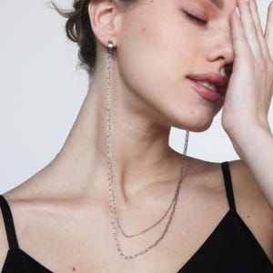 Peut inclure: Une boucle d'oreille en chaîne argentée avec un anneau en haut. La chaîne est longue et descend jusqu'au cou. La boucle d'oreille est portée par une personne dont la main couvre une partie du visage.