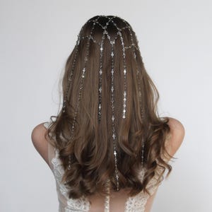 Può includere: Un copricapo argentato con un delicato design a catena. Il copricapo è indossato su lunghi capelli castani ondulati. Le catene scendono sulla schiena, adornate da piccoli abbellimenti rotondi. La persona indossa un top di pizzo bianco.