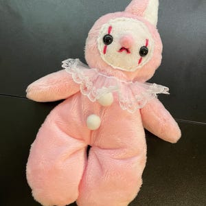 Könnte beinhalten: Eine Plüsch-Clownpuppe aus weichem rosa Stoff mit einem weißen Gesichtsbereich, schwarzen Knopfaugen und roten Stickereien. Die Puppe hat eine spitze Mütze, einen weißen Spitzenkragen mit Rüschen und zwei kleine weiße Bommeln auf der Brust.