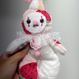Puede incluir: Un muñeco payaso de peluche con un patrón de fresas en tonos blanco y rosa. El muñeco tiene un cuello de encaje blanco, un pequeño pompón rosa en el gorro y el pecho, ojos de botón negros y marcas rojas cosidas bajo los ojos.