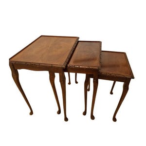 May include: A set of three antique nesting tables in a warm brown wood finish. Each table has a rectangular top with carved edges and elegant, curved legs. The tables are designed to fit neatly within each other, a classic design.