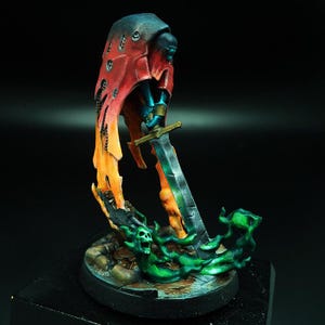 May include: A miniature figure of a cloaked figure with a sword, standing on a base with green and orange flames. The figure is painted in shades of blue, red, and orange.
