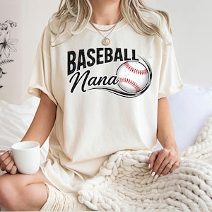 May include: A cream-coloured t-shirt featuring the text "Baseball Nana" in black script and a baseball graphic. The baseball is red and white with black stitching, and is encircled by black curved lines.