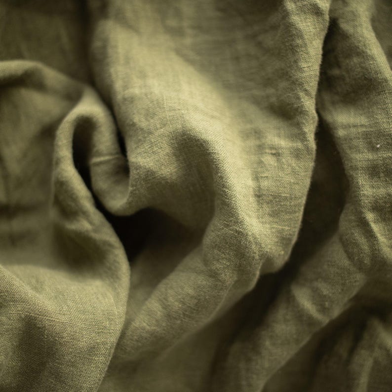May include: Close-up of olive green linen fabric with visible texture and folds. The fabric appears soft and slightly wrinkled, suggesting a natural fiber textile. The color is a muted, earthy tone.