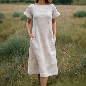 May include: A light beige linen dress with short sleeves and a boat neckline. The dress has a fitted bodice and a flared skirt with pockets. The model is standing in a field of tall grass.