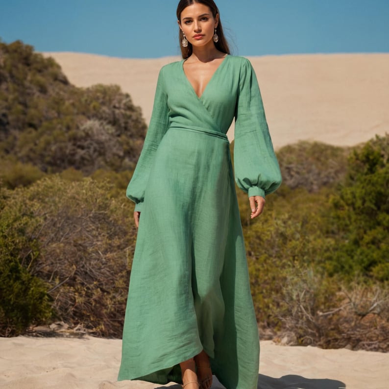 May include: A long, flowing, sage green wrap dress with a V-neckline and long, voluminous sleeves. The dress is cinched at the waist with a matching belt. The model is standing on a sandy beach with a natural background.