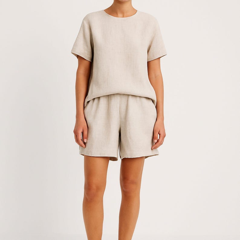 May include: A beige linen two-piece set. The set includes a short-sleeved, crew neck top and matching shorts. The shorts have an elastic waistband. The fabric has a slightly textured appearance.