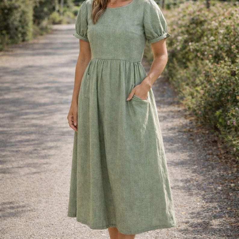 May include: A light green linen dress with short, puffed sleeves and a round neckline. The dress features a fitted bodice, an empire waist, and a flowing skirt with two front pockets. The dress falls to mid-calf length.