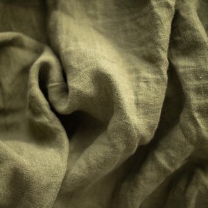 May include: Close-up of olive green linen fabric with soft folds and a textured surface. The fabric has a natural, slightly wrinkled appearance, suggesting a relaxed and comfortable textile.
