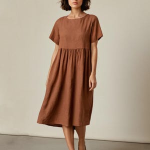 Op de afbeelding: Een bruine linnen jurk met korte mouwen en een gerimpelde taille. De jurk valt tot halverwege de kuit en heeft een ronde halslijn. De kleur is een warme, aardse tint, geschikt voor vrijetijdskleding.