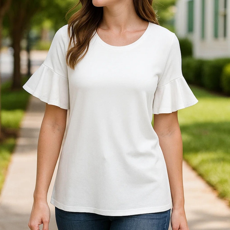 May include: A white, short-sleeved top with a round neckline and flared sleeves. The top is made of a soft fabric and is worn with blue jeans. The background includes a sidewalk, trees, and a building.